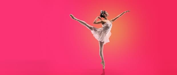 Image shows a single dancer spotlighted in orange against a vivid pink background. The dancer has dark hair pulled into a bun on the back of her head. She is wearing a white tiara and is wearing a fine white dress, with a mid-thigh-length skirt in diaphanous fabric and a sleeveless top with straps. The fine garland of grey flowers form the left shoulder strap and crosses her body to the right side of her waist. The dancer is facing to the right of the image, standing on point on her left leg, with her right leg raised in arabesque, the toe pointing to the top left of the picture. Her left arm is raised and outstretched towards the top right hand corner of the image. Her left arm is behind her with the elbow bent and her wrist bent so her fingers touch her shoulder