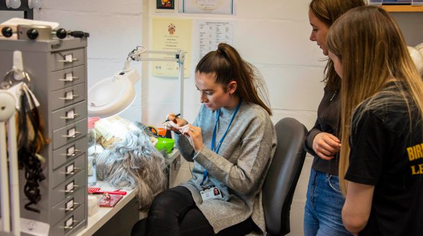 Members of BRB’s LEAP Ambassadors gather around a member of BRB’s technical department to watch a wig being constructed. There is a member of BRB’s technical team sat down on a grey swivel chair. The LEAP Ambassadors are standing behind the technical team member watching on as the team member is making a grey, long-haired wig. A white ring-light illuminates the wig which is attached to a mannequin head, laying on a desk which the technical team member is sat at. There are various posters on the white brick wall and a set of grey office drawers on the desk.