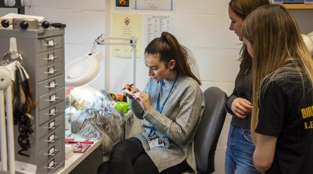 Members of BRB’s LEAP Ambassadors gather around a member of BRB’s technical department to watch a wig being constructed. There is a member of BRB’s technical team sat down on a grey swivel chair. The LEAP Ambassadors are standing behind the technical team member watching on as the team member is making a grey, long-haired wig. A white ring-light illuminates the wig which is attached to a mannequin head, laying on a desk which the technical team member is sat at. There are various posters on the white brick wall and a set of grey office drawers on the desk.