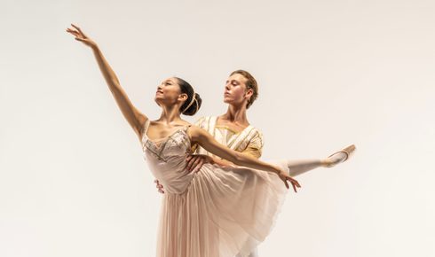 Maïlène Katoch and Alfie Shacklock in a photoshoot for BRB2. Alfie is holding Maïlène around the waist whilst Maïlène stands on pointe and reaches upwards. The couple are looking off to the left of the image. Maïlène has dark hair which is tied back and is wearing an elegant pink, knee-length dress, tights and pink pointe shoes. Alfie has short blonde hair and is wearing a white and gold top, tights, and white ballet flats.