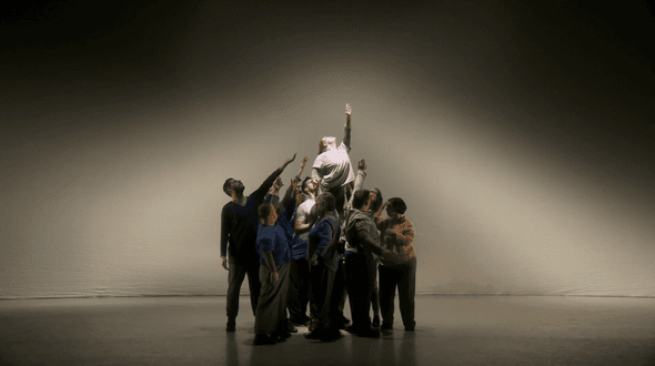 A group of dancers stand huddled in the centre of the stage. They are all pointing up to the sky. One of them has been lifted by the others above their heads and she is in a spotlight as she too reaches up.