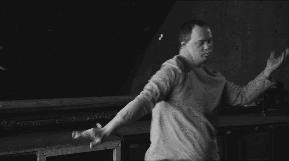This is a black and white image. Captured in a deserted bar or commercial kitchen. Viewed from the waist up, a lone dancer stands just the right of centre, his arms are out to both sides, and out of focus as he moves through his dance.