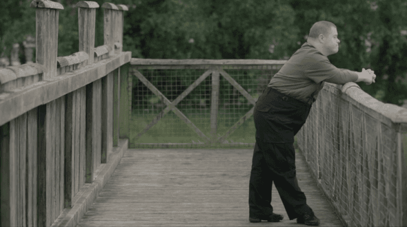 A lone figure, wearing black trousers and a long sleeved pale brown top stands to the right of the image leaning on a wooden rail and looking out of the image to the right. He seems to be standing on a wooden pathway or bridge as we can see more wood walls to the left, wooden planks on the floor, and int he distance a fence in front of green leaved trees.