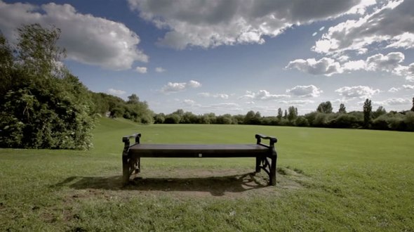 Bright summery day. Blue sky with lots of lively white clouds. Green field, hedges visible in the distance. Single, empty, park bench in foreground