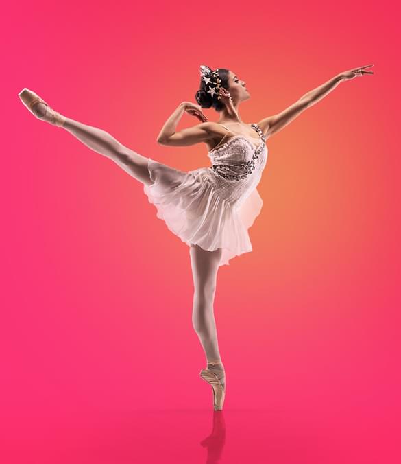 Image shows a single dancer spotlighted in orange against a vivid pink background. The dancer has dark hair pulled into a bun on the back of her head. She is wearing a white tiara and is wearing a fine white dress, with a mid-thigh-length skirt in diaphanous fabric and a sleeveless top with straps. The fine garland of grey flowers form the left shoulder strap and crosses her body to the right side of her waist. The dancer is facing to the right of the image, standing on point on her left leg, with her right leg raised in arabesque, the toe pointing to the top left of the picture. Her left arm is raised and outstretched towards the top right hand corner of the image. Her left arm is behind her with the elbow bent and her wrist bent so her fingers touch her shoulder