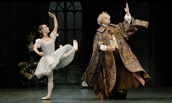 Two dancers stand side by side on stage. A young woman dressed in a pale blue dress stands with her left leg and arm raised stiffly, her right arm curved in front of her. A man, in long, richly patterned clock also raises his left hand and curves hiis right arm in front of him.