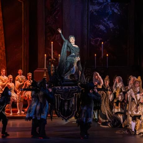 Daria Stanciulescu as Carabosse being carried in on a chariot in The Sleeping Beauty. Daria is lit by a spotlight and rises high above the onlooking crowd as Carabosse’s minions carry in a large, black chariot above their heads. Daria is raising the right arm and is looking out at the audience. Daria is wearing an opulent black gown with large headpiece. In the background, a golden set lit by candelabras and warm stage lights encapsulates the scene.