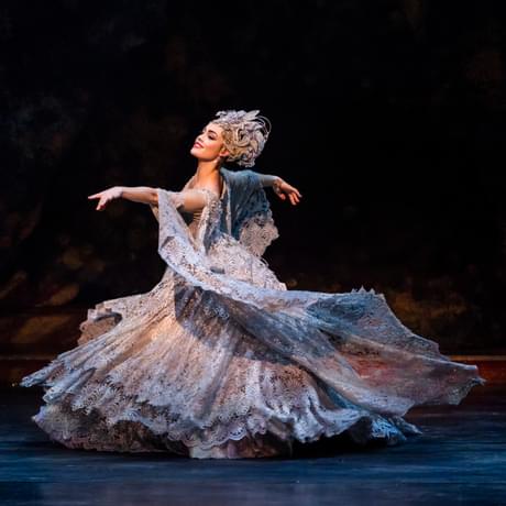 Eilis Small as the Lilac Fairy in The Sleeping Beauty. Eilis has dark hair and is wearing an opulent lilac gown made of detailed lace with a floral pattern and a headpiece made of flowers and complete with light purple jewels. Eilis is smiling and is leading with the right arm, left arm trailing behind as Eilis gracefully moves across the warmly-lit stage.