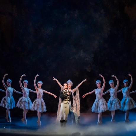 Tristram Kenton of Lachlan Monaghan as Prince Florimund in The Sleeping Beauty. A spotlight shines on Lachlan and Lachlan is standing centrally on a stage upon which fog is rolling across. Eilis Small as The Lilac Fairy is standing behind Lachlan with arms raised. Other dancers in a line either side of Lachlan, standing en pointe with arms raised. Lachlan is looking out to the audience, with the right arm across the chest. Lachlan has short dark hair and is wearing a black solider’s jacket which has gold detailing, a white shirt with black neck tie, white fitted trousers and black knee-high boots.