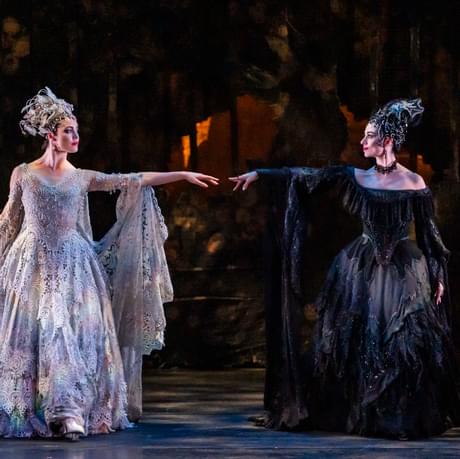 Daria Stanciulescu as Carabosse and Eilis Small as the Lilac Fairy in The Sleeping Beauty. These two sisters and sworn enemies are looking one another in the eye and are reaching out to each other. Daria is wearing an opulent black gown with large headpiece. Eilis is wearing an equally opulent lilac dress and headpiece. The pair stand out against a gloomy backdrop with hints of gold peeking through a curtain.