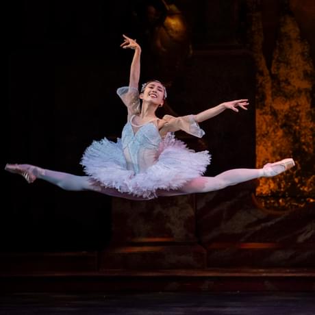 Tristram Kenton of Yu Kurihara as Princess Aurora in The Sleeping Beauty. Yu is performing a split jeté (both legs split whilst in a jump, one arm raised and one out to the side) on a stage with a golden, glowing backdrop. Yu has dark hair which is tied back and secured using an elegant tiara. Yu is wearing a light pink tutu which has sequinned details and opaque 1/4 length sleeves, pink tights and pink pointe shoes.