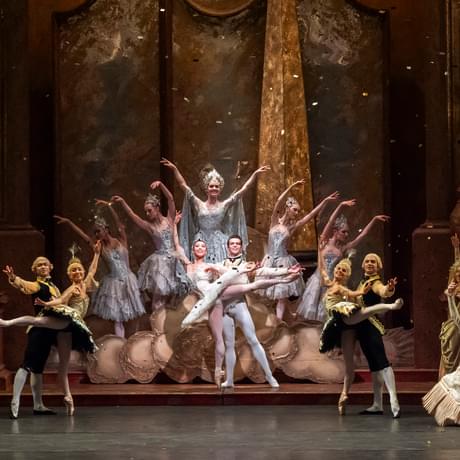 Tristram Kenton of Lachlan Monaghan as Prince Florimund and Yu Kurihara as Princess Aurora standing amongst the cast of The Sleeping Beauty as golden glitter rains down from the ceiling. The entire cast are kneeling or standing in various poses with one arm raised and the other lowered (lower arm towards the audience). The cast are wearing extravagant period outfits, complete with wigs, tutus and headpieces. A small group of dancers, including the Lilac Fairy, Prince Florimund and Princess Aurora are standing on a raised platform in the centre of the stage. Lachlan is supporting Yu who is standing en pointe. The detailed golden set frames the group of dancers.