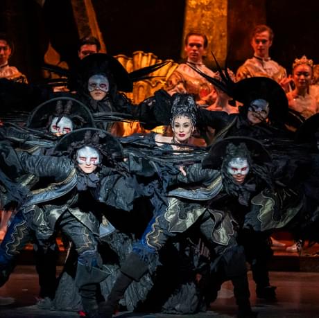 Tristram Kenton of Daria Stanciulescu as Carabosse with her minions around her in The Sleeping Beauty. The group are all leaning to the right of the image. Daria smiles subtly as her minions, all dressed in black and wearing eye masks surround her. Daria is wearing an opulent black gown with large headpiece. Anxious onlookers watch from the rear of the stage as the group dance in front of them.