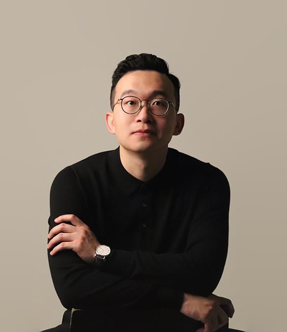 A professional headshot of Yi Wei looking at the camera against a pastel grey background. Yi Wei is sitting with arms folded, wearing a black jumper, round black glasses and a watch with a white watch face and black strap. Yi Wei has a neutral expression on their face.