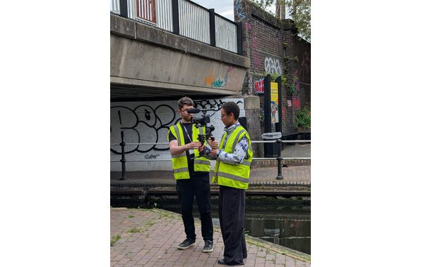 A young woman and a young man are holding a black film camera. They are looking down at it as the young man explains something. Both are wearing bright yellow high-vis jackets and dark trousers. They are standing on one side of a canal with a bridge behind them.