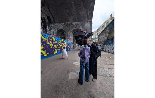 Two young women are standing with their backs to the camera in an industrial open air space. One of them is reaching up to set up some lighting equipment. To their right is a ballet dancer on the points of her feet next to a graffitied wall.