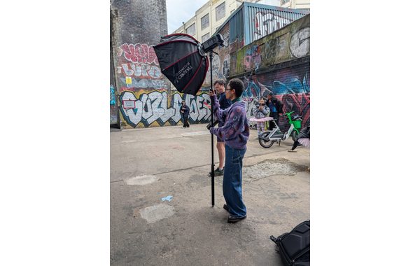A young woman is holding the pole of some black lighting equipment. She is standing side-on to the camera. She is wearing a purple cardigan and dark trousers. Behind her are graffitied walls.