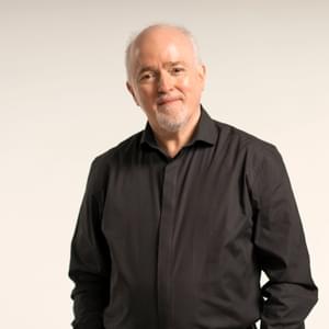 Paul Murphy, Principal Conductor of the Royal Ballet Sinfonia is standing against an off-white backdrop, smiling for a headshot. Paul is facing forwards, looking at the camera and smiles with mouth closed. Paul has grey/white short hair and a trimmed goatee. Paul is wearing a black formal shirt and black trousers.
