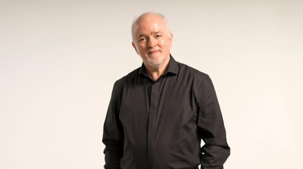 Paul Murphy, Principal Conductor of the Royal Ballet Sinfonia is standing against an off-white backdrop, smiling for a headshot. Paul is facing forwards, looking at the camera and smiles with mouth closed. Paul has grey/white short hair and a trimmed goatee. Paul is wearing a black formal shirt and black trousers.