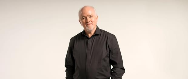 Paul Murphy, Principal Conductor of the Royal Ballet Sinfonia is standing against an off-white backdrop, smiling for a headshot. Paul is facing forwards, looking at the camera and smiles with mouth closed. Paul has grey/white short hair and a trimmed goatee. Paul is wearing a black formal shirt and black trousers.