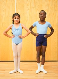 A young girl and boy stand next to each other, hands on their hips. She is wearing a pale blue leotard with pink tights and satin ballet shoes. He is wearing a pale blue leotard with navy shorts, white socks and white ballet shoes.