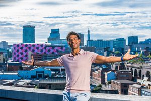 Carlos Acosta is seen sitting on a wall, arms stretched wide and smiling, with a panorama of the Birmingham skyline behind him