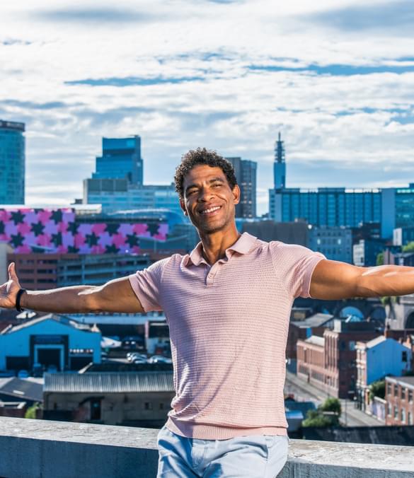 Carlos Acosta is seen sitting on a wall, arms stretched wide and smiling, with a panorama of the Birmingham skyline behind him