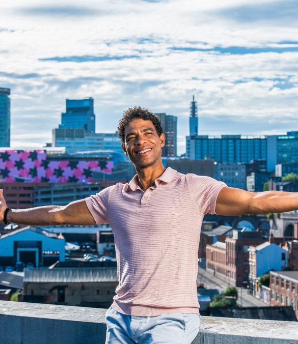 Carlos Acosta is seen sitting on a wall, arms stretched wide and smiling, with a panorama of the Birmingham skyline behind him