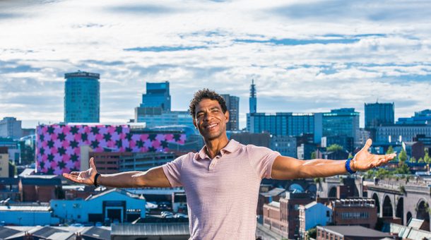 Carlos Acosta is seen sitting on a wall, arms stretched wide and smiling, with a panorama of the Birmingham skyline behind him