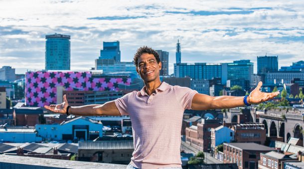 Carlos Acosta is seen sitting on a wall, arms stretched wide and smiling, with a panorama of the Birmingham skyline behind him