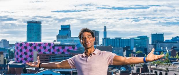 Carlos Acosta is seen sitting on a wall, arms stretched wide and smiling, with a panorama of the Birmingham skyline behind him