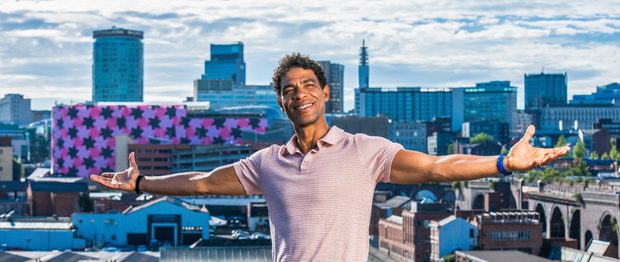 Carlos Acosta is seen sitting on a wall, arms stretched wide and smiling, with a panorama of the Birmingham skyline behind him