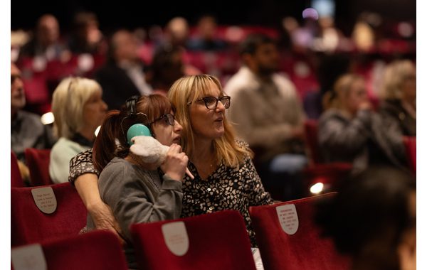 This image is in a theatre auditorium during a relaxed performance. In the central foreground are two women seated with other audience members around them. There are empty seats next to them and in front of them. The younger woman on the left has ear defenders and is holding a fluffy toy while the other lady has their arm around them. Both are looking to the right engrossed with what's happening on stage.