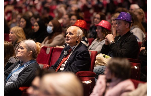 This image shows a theatre audience. In the centre is an older man wearing a headset during an audio-described performance. Behind him are theatre volunteers wearing sparkly hats.