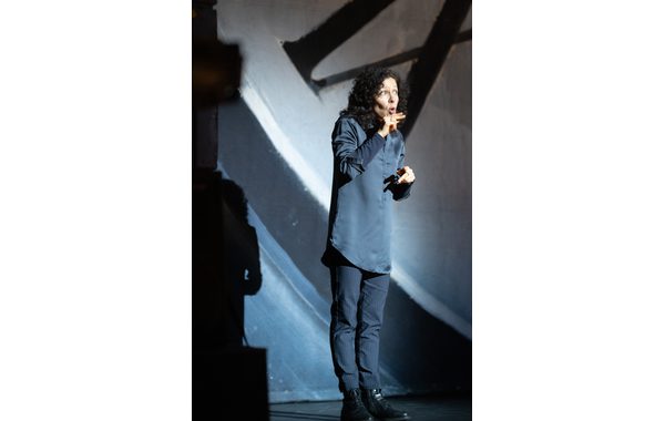This image shows a lady signing what's happening on stage. She is looking surprised and has both her hands with her fingers held out straight. In the background is some roman numerals which form part of the large clock making up some of the staging.