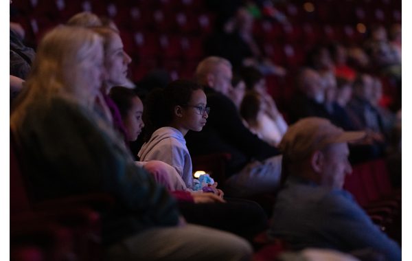 The image shows a full audience in a theatre. Most of them are out of focus but in the centre is a little girl in glasses. She is holding a fidget toy and is leaning forward slightly looking at what's happening on stage.