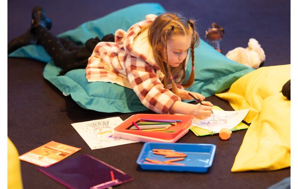 An image of a little girl lying on a beanbag in the theatre foyer. She is colouring in an image as part of what we offer during a relaxed performance. There are trays of crayons around her. She is concentrating on what she's colouring.