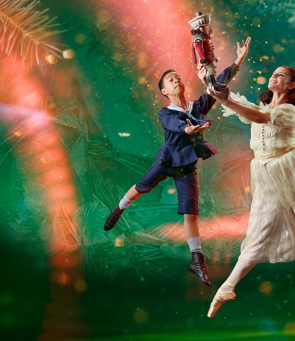 There are two dancers, slightly to the right of centre. The background is dark green with a large, brighter spotlight that doesn't reach the bottom corners of the image. In the background are giant pine needles, a scattering of small lighter areas, and curved beams of red light, that look paler when they are within the spotlight. The dancer in the foreground has shoulder length brown hair tied back with a white ribbon. She is representing the character of Clara. She is facing towards the left of the picture. She wears a white dress and a pair of off-white ballet pointe shoes. She is in mid leap with her left leg stretched out behind her. Her right leg is pointing towards the floor. She's holding up in front of her with both hands a Nutcracker doll, painted to look like a soldier in a red jacket and white trousers. The dancer behind Clara is representing the character of Fritz, Clara's younger brother. He has short dark hair and is wearing a dark blue sailor suit with britches to just below his knee. On his feet are white socks and soft black ballet shoes. He is facing Clara and his arms are raised as if trying to steal the Nutcracker doll from her. Photo copyright Manvir Rai.