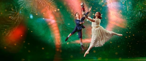 There are two dancers, slightly to the right of centre. The background is dark green with a large, brighter spotlight that doesn't reach the bottom corners of the image. In the background are giant pine needles, a scattering of small lighter areas, and curved beams of red light, that look paler when they are within the spotlight. The dancer in the foreground has shoulder length brown hair tied back with a white ribbon. She is representing the character of Clara. She is facing towards the left of the picture. She wears a white dress and a pair of off-white ballet pointe shoes. She is in mid leap with her left leg stretched out behind her. Her right leg is pointing towards the floor. She's holding up in front of her with both hands a Nutcracker doll, painted to look like a soldier in a red jacket and white trousers. The dancer behind Clara is representing the character of Fritz, Clara's younger brother. He has short dark hair and is wearing a dark blue sailor suit with britches to just below his knee. On his feet are white socks and soft black ballet shoes. He is facing Clara and his arms are raised as if trying to steal the Nutcracker doll from her. Photo copyright Manvir Rai.