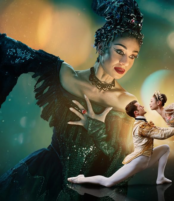 Two dancers are in the foreground looking at each other with a much larger figure in the background towering over them. The background is a blended blue and green at the top of the image. The image fades to black at the bottom. Surrounding the two dancers in the foreground is a glowing gold light with spots and streams of light surrounding the centre of the image. This gold is reflected onto the towering figure’s face. The two dancers in the foreground are towards the right of the image and are playing the characters of the Prince and Sleeping Beauty. The dancer playing the Prince has brown, short hair, is wearing white, fitted tights, white ballet flats and a cream jacket with gold detailing. He is on the left of the couple, kneeling one knee, looking up at Sleeping Beauty, with arms extended, holding her waist. The dancer playing Sleeping Beauty has dark hair in a bun, is wearing a white tutu with silver detailing, a sequinned top, sparkling tiara and is on pointe on her right leg, with her left leg extended pointing directly upwards. The figure in the background is playing Carabosse, she is positioned to the right of the foreground dancers, and is looking down at them. The dancer playing Carabosse has black hair with a silver headpiece, is wearing a gothic black dress with sequinned detailing and has black eye-makeup and red lipstick. Carabosse has her left arm clutching at her chest, a ring with large, red gemstone is on her middle finger, her right arm is extended towards the upper left and a dramatic black sleeve drapes towards the floor.