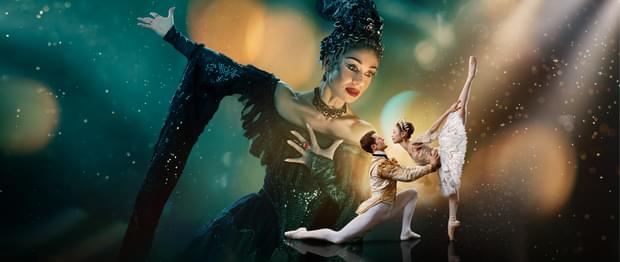 Two dancers are in the foreground looking at each other with a much larger figure in the background towering over them. The background is a blended blue and green at the top of the image. The image fades to black at the bottom. Surrounding the two dancers in the foreground is a glowing gold light with spots and streams of light surrounding the centre of the image. This gold is reflected onto the towering figure’s face.   The two dancers in the foreground are towards the right of the image and are playing the characters of the Prince and Sleeping Beauty. The dancer playing the Prince has brown, short hair, is wearing white, fitted tights, white ballet flats and a cream jacket with gold detailing. He is on the left of the couple, kneeling one knee, looking up at Sleeping Beauty, with arms extended, holding her waist. The dancer playing Sleeping Beauty has dark hair in a bun, is wearing a white tutu with silver detailing, a sequinned top, sparkling tiara and is on pointe on her right leg, with her left leg extended pointing directly upwards.   The figure in the background is playing Carabosse, she is positioned to the right of the foreground dancers, and is looking down at them. The dancer playing Carabosse has black hair with a silver headpiece, is wearing a gothic black dress with sequinned detailing and has black eye-makeup and red lipstick. Carabosse has her left arm clutching at her chest, a ring with large, red gemstone is on her middle finger, her right arm is extended towards the upper left and a dramatic black sleeve drapes towards the floor.