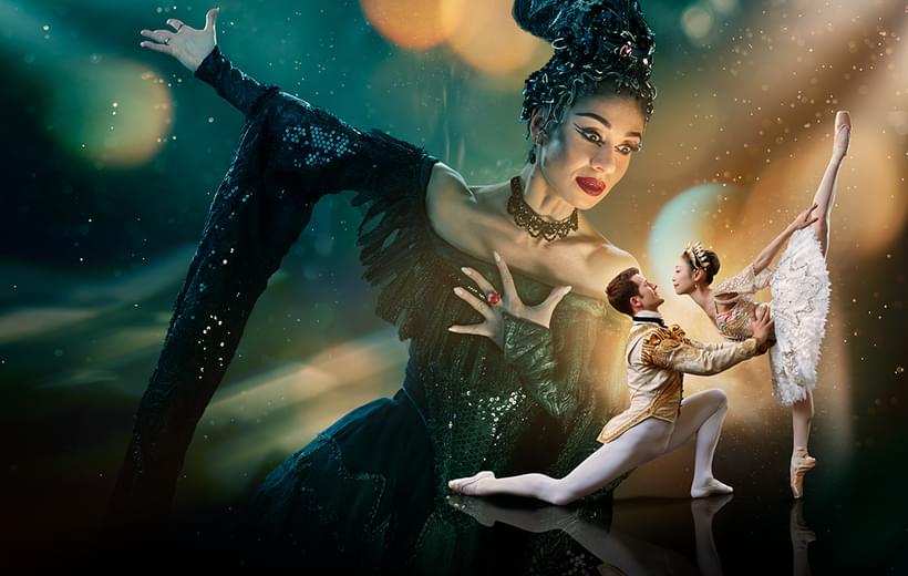 Two dancers are in the foreground looking at each other with a much larger figure in the background towering over them. The background is a blended blue and green at the top of the image. The image fades to black at the bottom. Surrounding the two dancers in the foreground is a glowing gold light with spots and streams of light surrounding the centre of the image. This gold is reflected onto the towering figure’s face.   The two dancers in the foreground are towards the right of the image and are playing the characters of the Prince and Sleeping Beauty. The dancer playing the Prince has brown, short hair, is wearing white, fitted tights, white ballet flats and a cream jacket with gold detailing. He is on the left of the couple, kneeling one knee, looking up at Sleeping Beauty, with arms extended, holding her waist. The dancer playing Sleeping Beauty has dark hair in a bun, is wearing a white tutu with silver detailing, a sequinned top, sparkling tiara and is on pointe on her right leg, with her left leg extended pointing directly upwards.   The figure in the background is playing Carabosse, she is positioned to the right of the foreground dancers, and is looking down at them. The dancer playing Carabosse has black hair with a silver headpiece, is wearing a gothic black dress with sequinned detailing and has black eye-makeup and red lipstick. Carabosse has her left arm clutching at her chest, a ring with large, red gemstone is on her middle finger, her right arm is extended towards the upper left and a dramatic black sleeve drapes towards the floor.