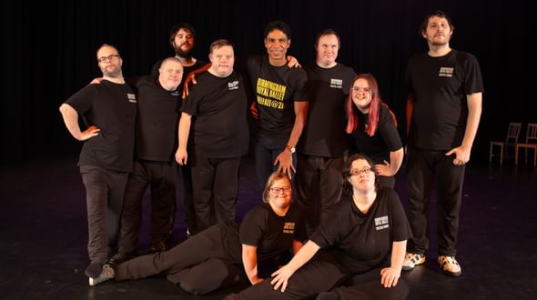 Members of Freefall Dance Company pose for a photo with BRB Director Carlos Acosta in the Patrick Studio. Seven members of Freefall are standing either side of Carlos with arms around each other. The other two members of the company are sitting on the floor with their legs off to one side. The group are smiling and are wearing matching black t-shirts with ‘Birmingham Royal Ballet, Freefall Dance’ written on the top left of the t-shirts. Carlos is wearing a black t-shirt with ‘Birmingham Royal Ballet, Freefall @ 21’ written in yellow across the front. The background is a black, cloth backdrop, the floor is black vinyl with some scuffs on it and there are two white chairs off to the right side. Credit John Palmer.