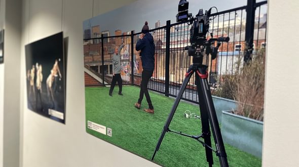 Henry McNab of the Freefall @ 21 exhibition at Birmingham Hippodrome. Dancers of the Freefall Dance Company are pictured in two different pictures hung against a grey wall. In the focal image, one of the dancers is amongst 5 white chairs in a green field surrounded with mature trees at the edges. The dancer is facing the camera with arms are spread outwards, and they are looking to the right of the image. There is more information in the bottom right corner. The image to the left of the focal image is out of focus, but pictures a group of the Freefall dancers wearing black in a dance studio. Credit: Henry McNab