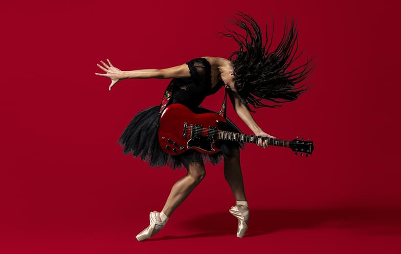 BRB Dancer Sofia Liñares is standing in the centre of the image, against a deep red background on which her shadow can be seen. Sofia Liñares is a dancer with long dark hair and is wearing a black sleeveless top, with black tutu, and pale pink pointe shoes. Sofia is standing with her left foot on pointe, her right leg is slightly bent at the knee, the top of her foot is resting on the ground. Sofia’s head is mid-motion, facing towards the ground, her hair is flying forwards with the strands of hair in an arch around her head. Sofia is holding a red and black electric guitar which is secured over her shoulder using a strap decorated with red roses and green leaves. Sofia’s right arm is facing the left as if she had just strummed the guitar, her left is holding the fret board in a chord. Image copyright Perou.
