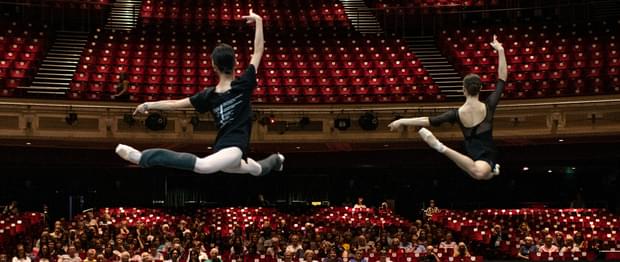 Looking from the back of the stage towards the auditorium, two dancers are leaping on the stage watched by an audience seated in the auditorium