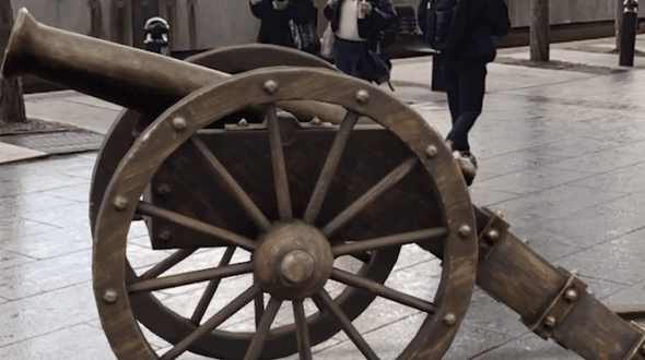 The cannon from the fight scene in The Nutcracker outside of BCU’s Parkside Building. The faux wooden cannon is in augmented reality on grey paving slabs, behind which three people are walking. The end of the cannon is pointing towards the left, and the large wheels are centred to the image.