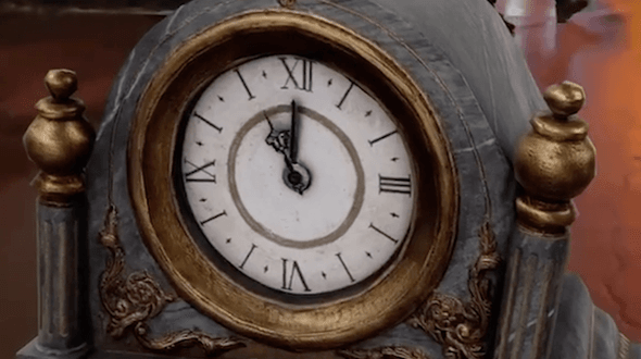 The clock from The Nutcracker at Hockley Social Club. The mantle clock is blue with gold detailing and a white face. The clock is taking up the majority of the frame, with a person’s feet visible in the background. The clock is ornate with decorative swirls on the front and the time in Roman numerals around the edge. The background is a orange/red due to lighting.