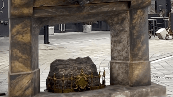 The fireplace from The Nutcracker in The Custard Factory Digbeth. The fireplace is a dark, beige stone with an ornate golden fire grate in the centre with faux coals. In the background there is grey paving and a black wall with a set of double doors. Off to the right there is a seating area.