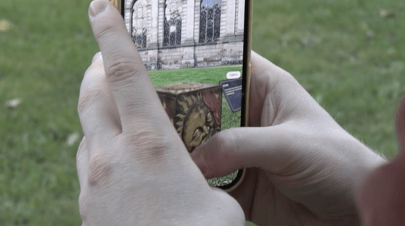 The Jack-in-the-Box in augmented reality on a phone. The phone has a yellow case and is being held with two hands. The Jack-in-the-Box can be seen on-screen, with a blue information card next to it. In the background, there is a park with grass. The Jack-in-the-Box has a golden sun on the front and a red piece of cloth draped over the front.